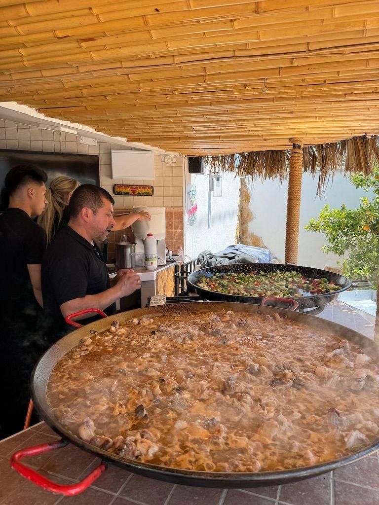 Cocineros cocinando paella en una gran sartén al aire libre bajo un techo de bambú, con vapor elevándose y otros ingredientes cerca.