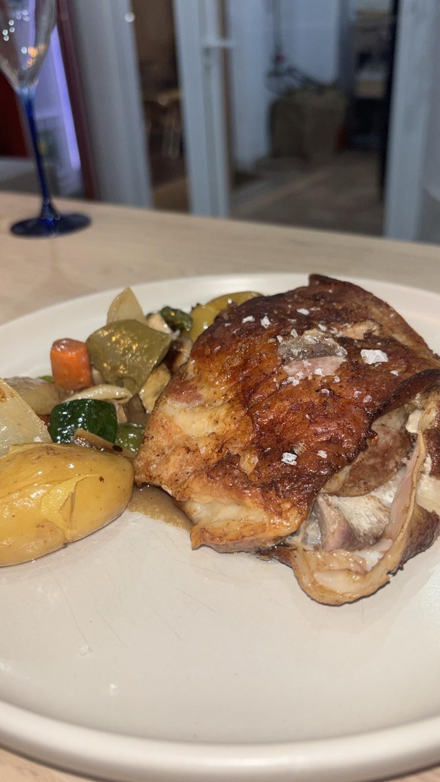 Muslo de pollo asado y verduras en un plato blanco, con una copa de vino de fondo.