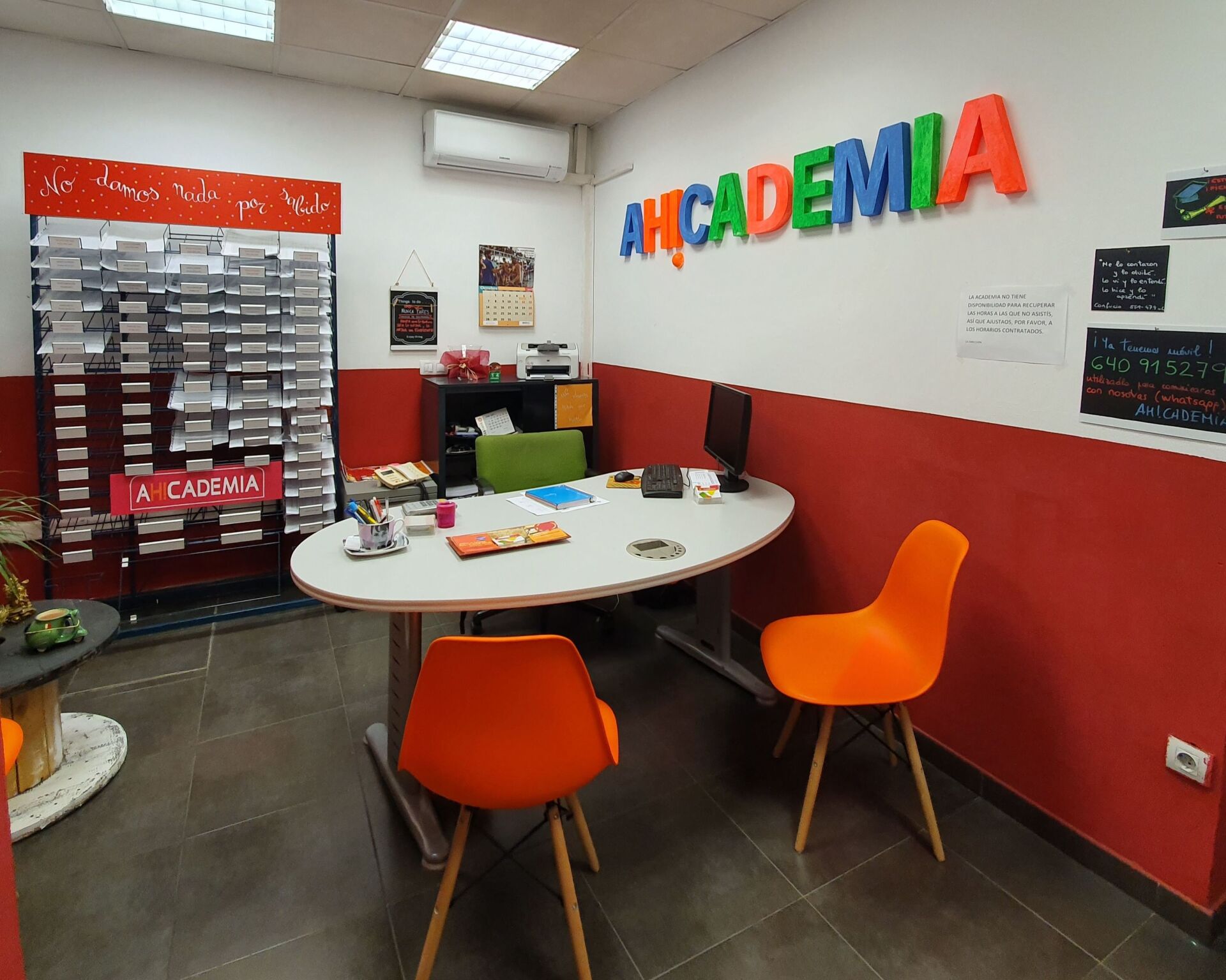Interior de una oficina con un escritorio y sillas naranjas. La pared tiene la colorida inscripción