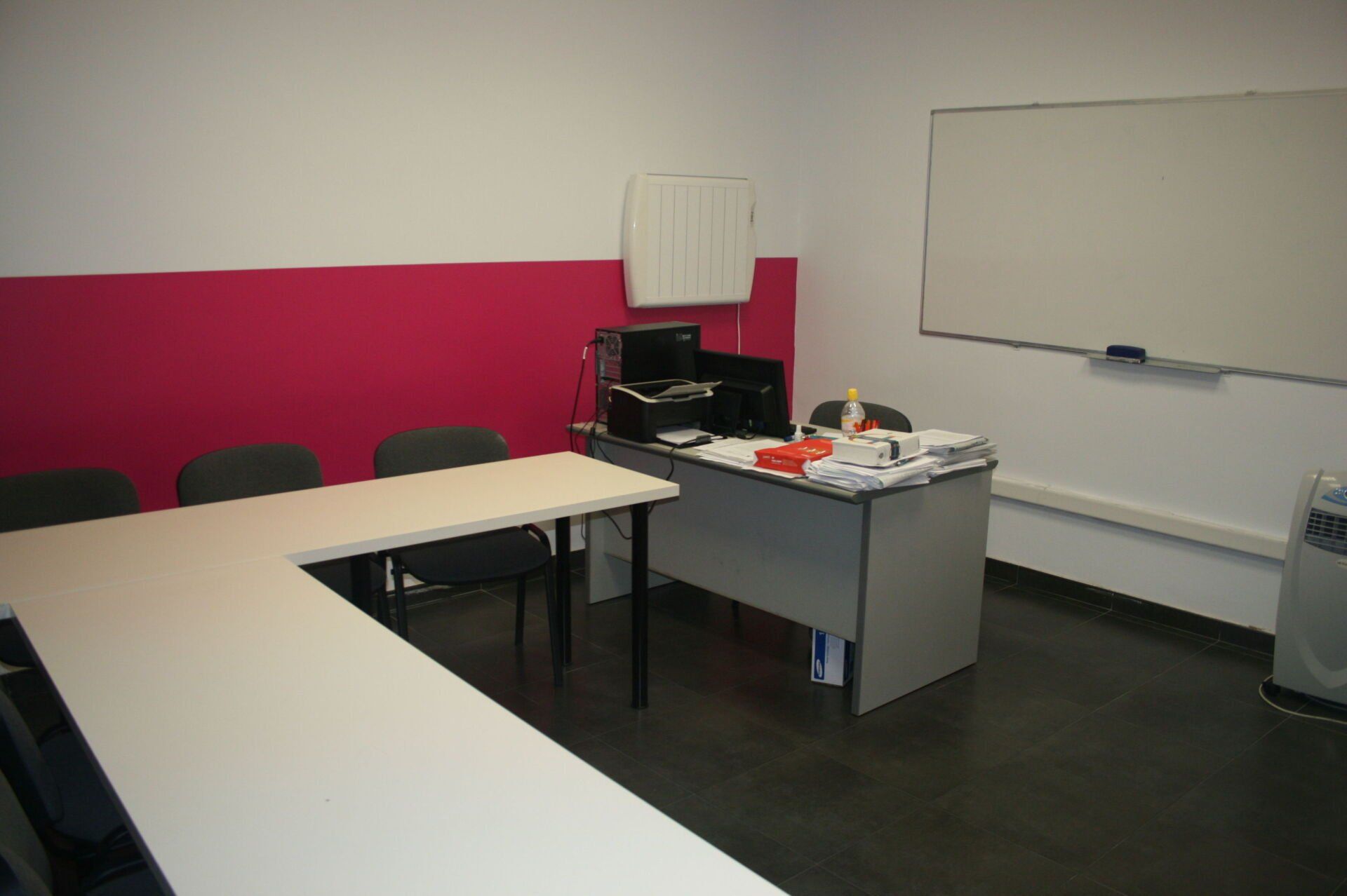 Mesa y escritorio en forma de L en una habitación con paredes rosas y blancas. También hay una pizarra y un ordenador.