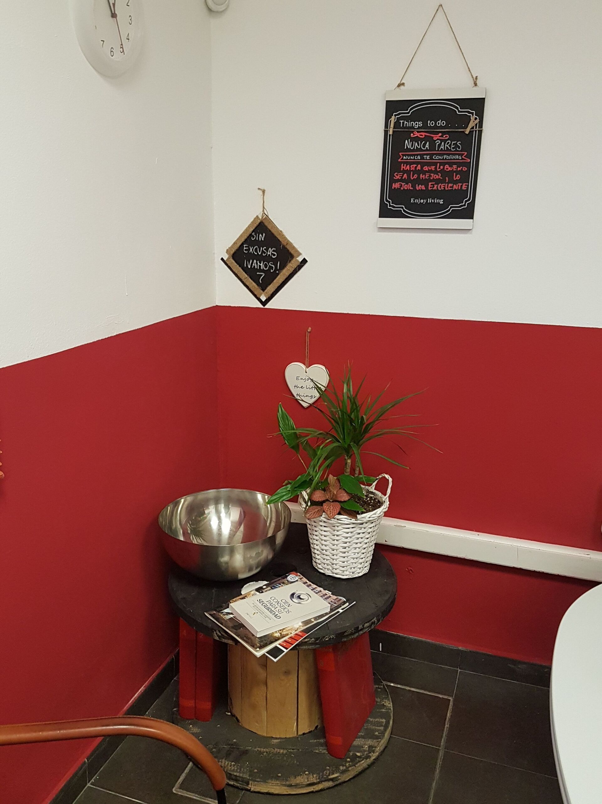 Rincón con mesa redonda, cuenco de metal, plantas y paredes rojas y blancas. Letrero colgado encima.