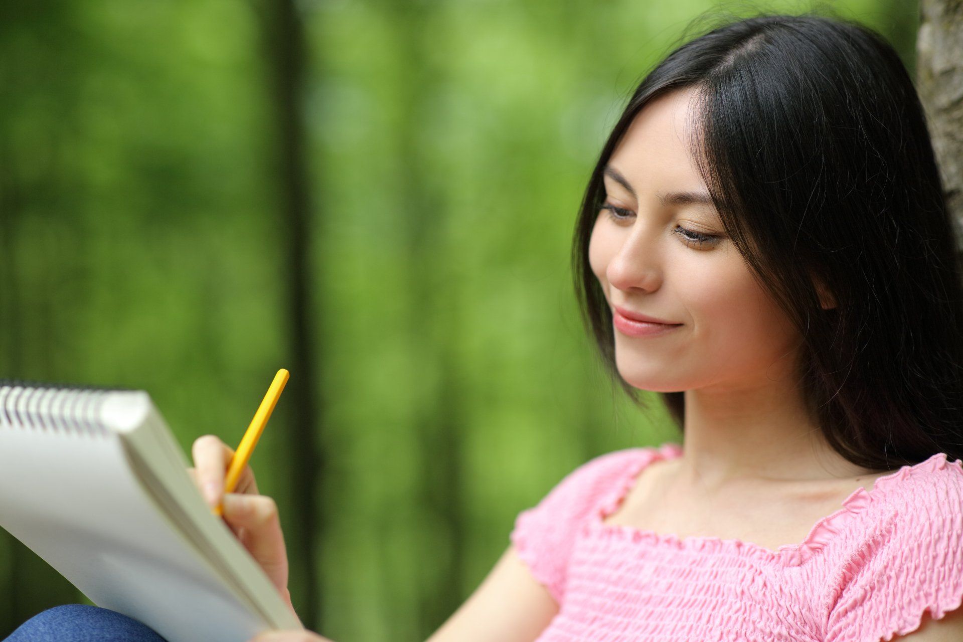 Mujer dibujando en un cuaderno con un lápiz en un bosque, sonriendo, vistiendo una blusa rosa.