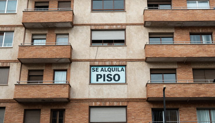 Fachada de un edificio de apartamentos con un cartel de «Se alquila piso» en la ventana central.