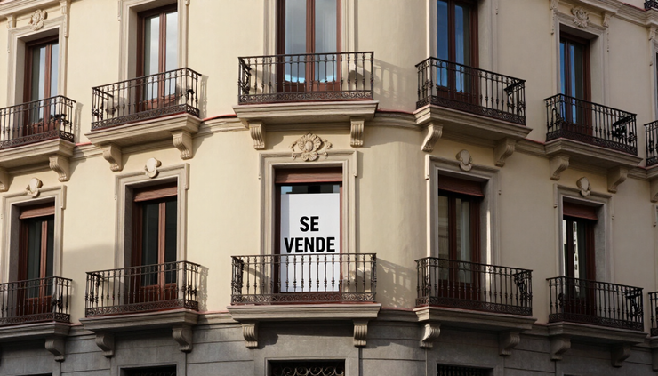 Edificio de apartamentos en esquina, con balcones y un cartel en la ventana central que dice «SE VENDE» (en venta)