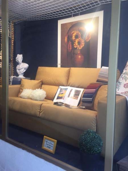 Sofá amarillo con cojines, arte, libros y un busto en exposición. Pared oscura, una pequeña planta y muestras de tela visibles.