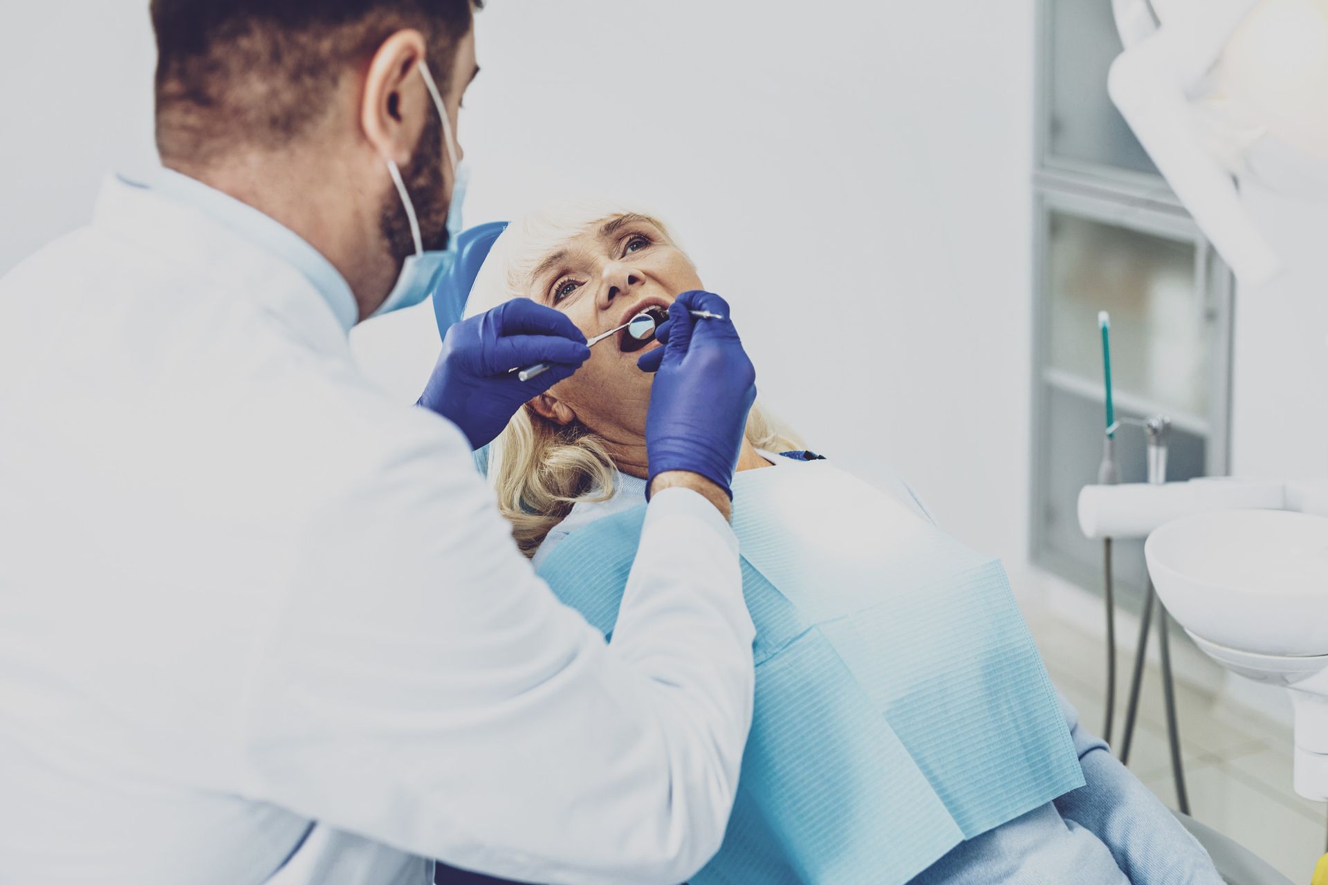 Dentista examinando los dientes de una mujer mayor; ambos usan máscaras y guantes en un consultorio dental.