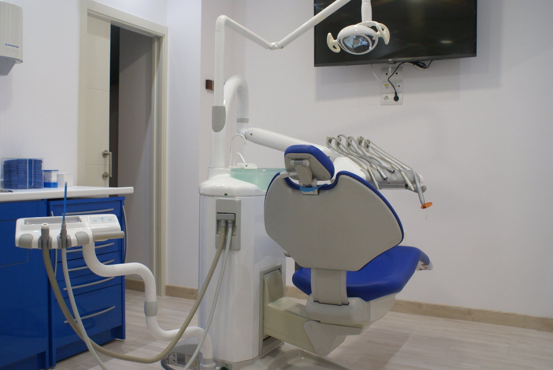 Sillón dental en sala de clínica; equipo azul y blanco; lámpara de techo; televisor montado en la pared.