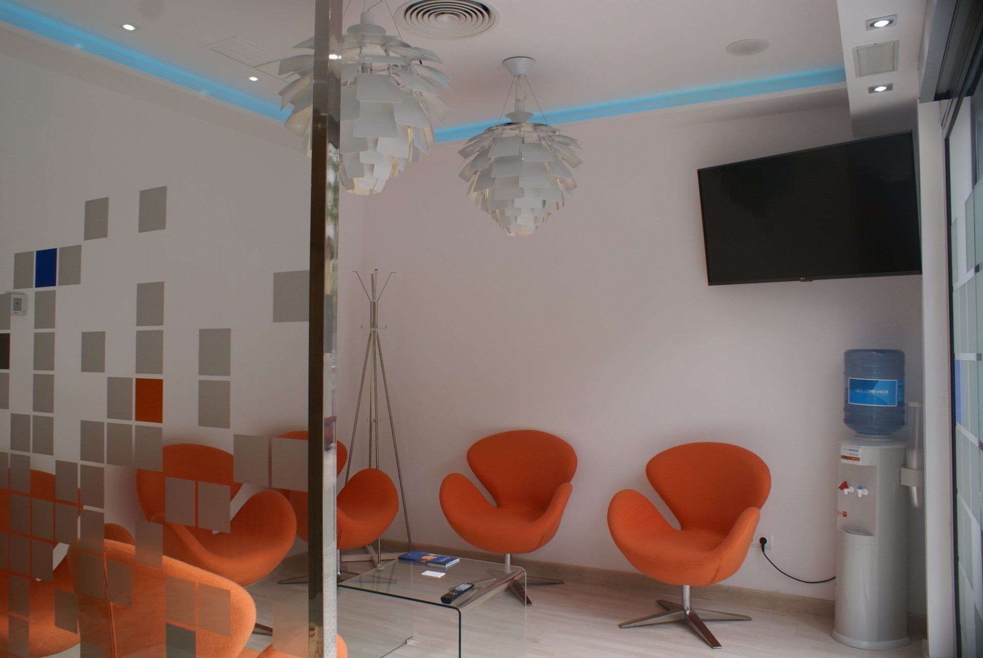 Sala de espera con sillas naranjas, TV, dispensador de agua y diseño de pared abstracto.