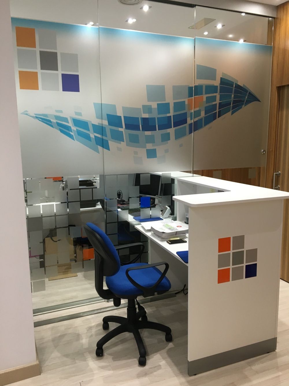 Mostrador de recepción con silla azul; pared de vidrio con cuadrados abstractos azules, naranjas y grises; entorno de oficina.