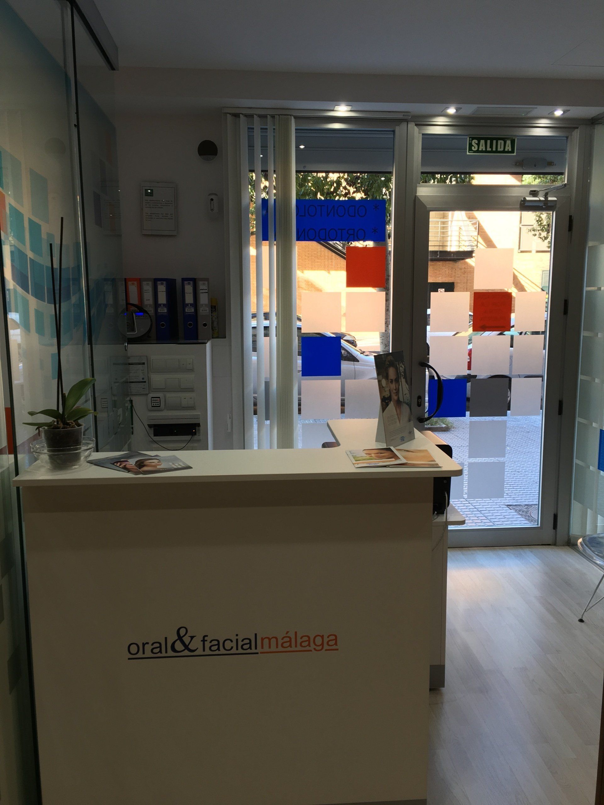 Zona de recepción de una clínica dental, con un escritorio blanco, una ventana con cuadrados de colores y una puerta de entrada.
