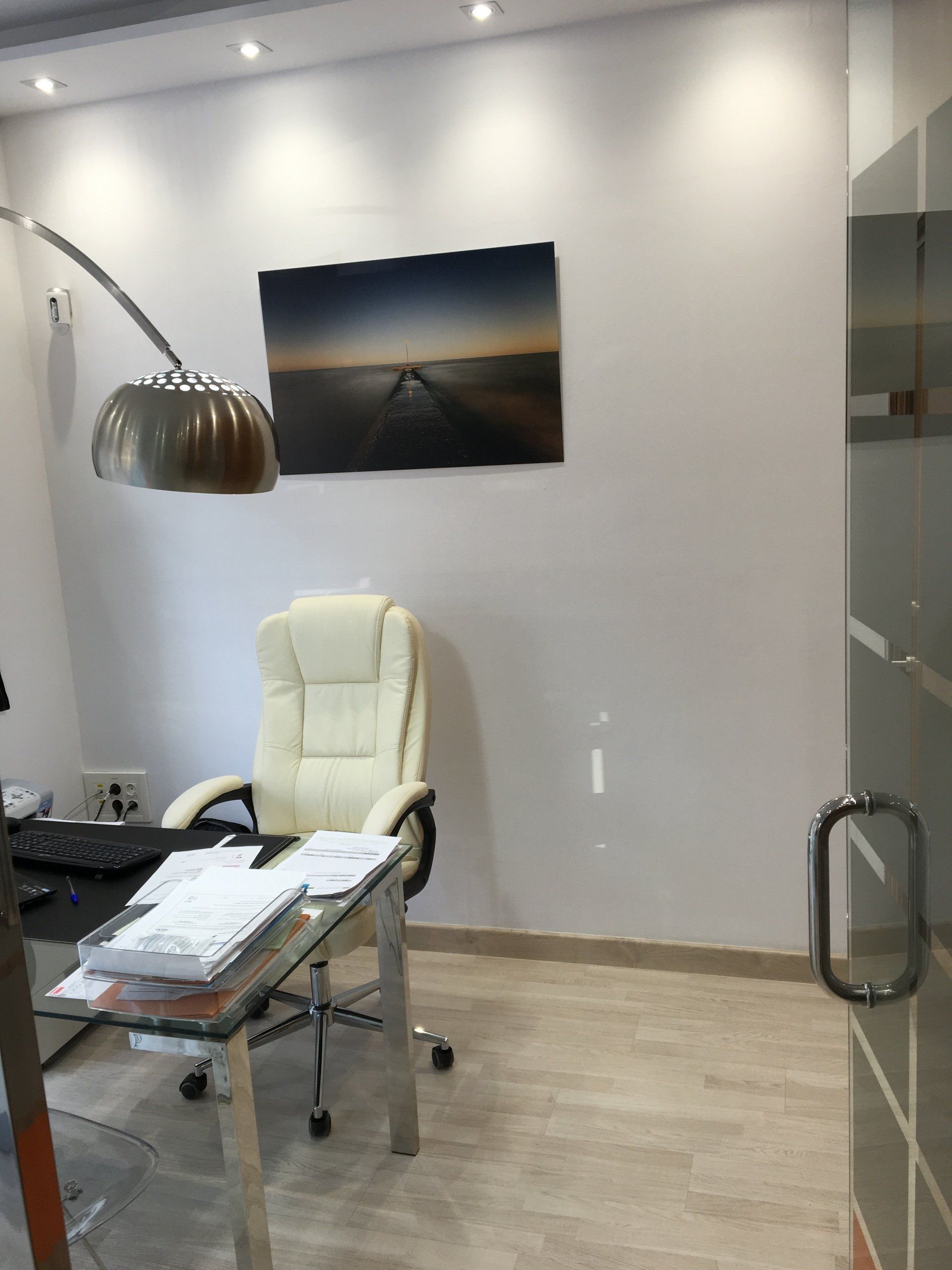 Oficina con pared blanca, escritorio, silla, cuadro y lámpara arqueada. El suelo es marrón claro.