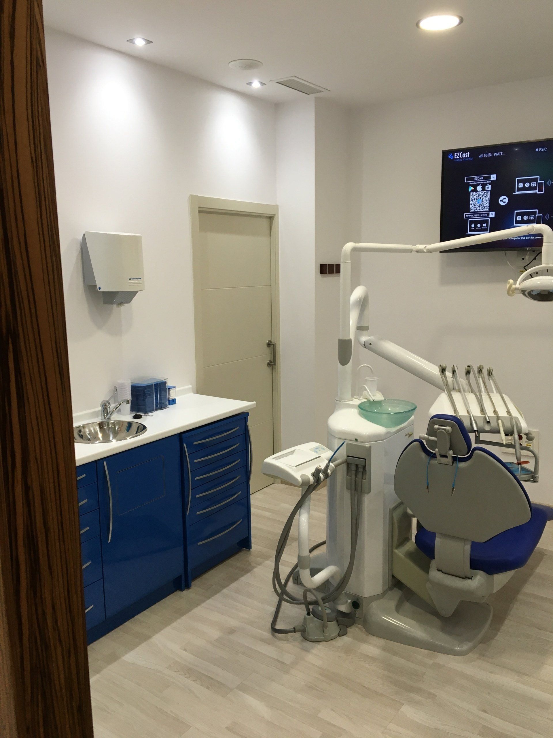 Consultorio dental: gabinetes azules, sillón dental, equipo, paredes blancas y una puerta.