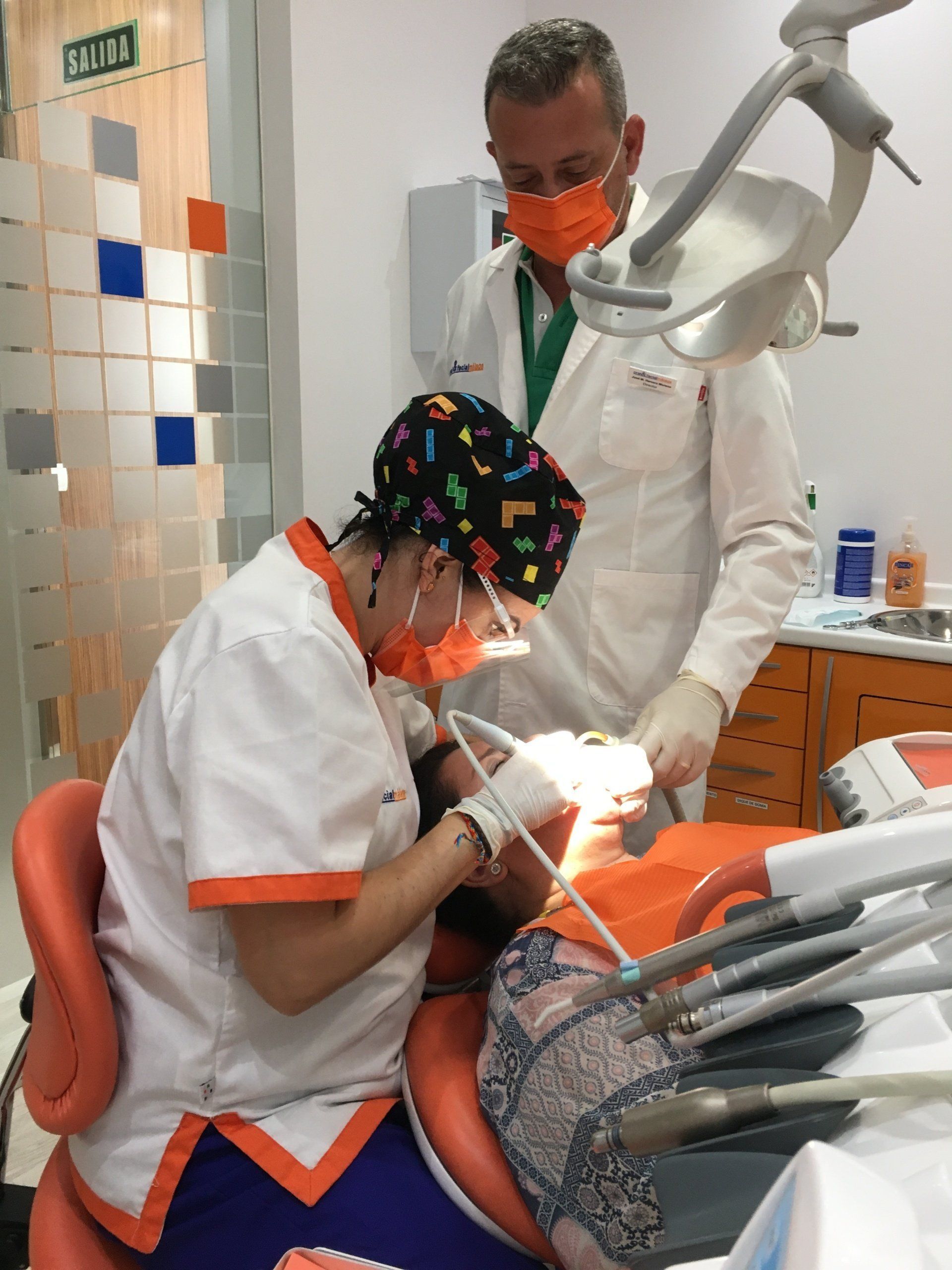 Dentista y asistente trabajando en los dientes de un paciente en una clínica dental. Ambos usan mascarillas y guantes.