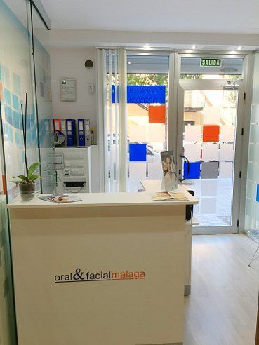 Mostrador de recepción de una clínica dental; logotipo de Oral & Facial Málaga. Puerta con cuadrados de colores; planta.