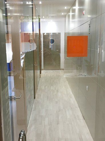 Pasillo con suelo de imitación madera clara y paredes de cristal, que da a una puerta metálica con ojo de buey. Cuadrado naranja en la pared.