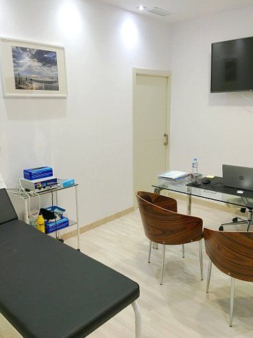 Consultorio médico: mesa de exploración, escritorio con computadora, dos sillas, cuadro en la pared y una puerta.