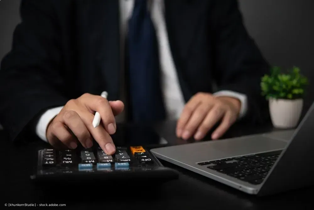Eine Person im Businessanzug benutzt an einem Schreibtisch einen Taschenrechner und einen Laptop; 