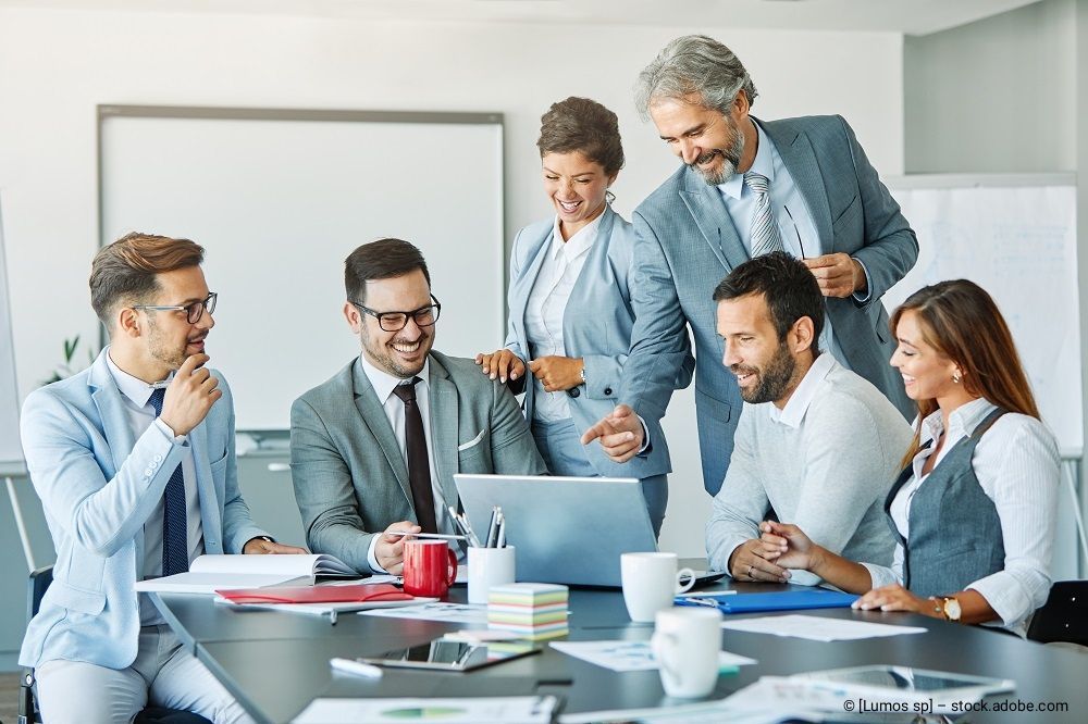 Eine bunte Gruppe von Geschäftsleuten sitzt und steht während einer gemeinsamen Besprechung um einen Konferenztisch.