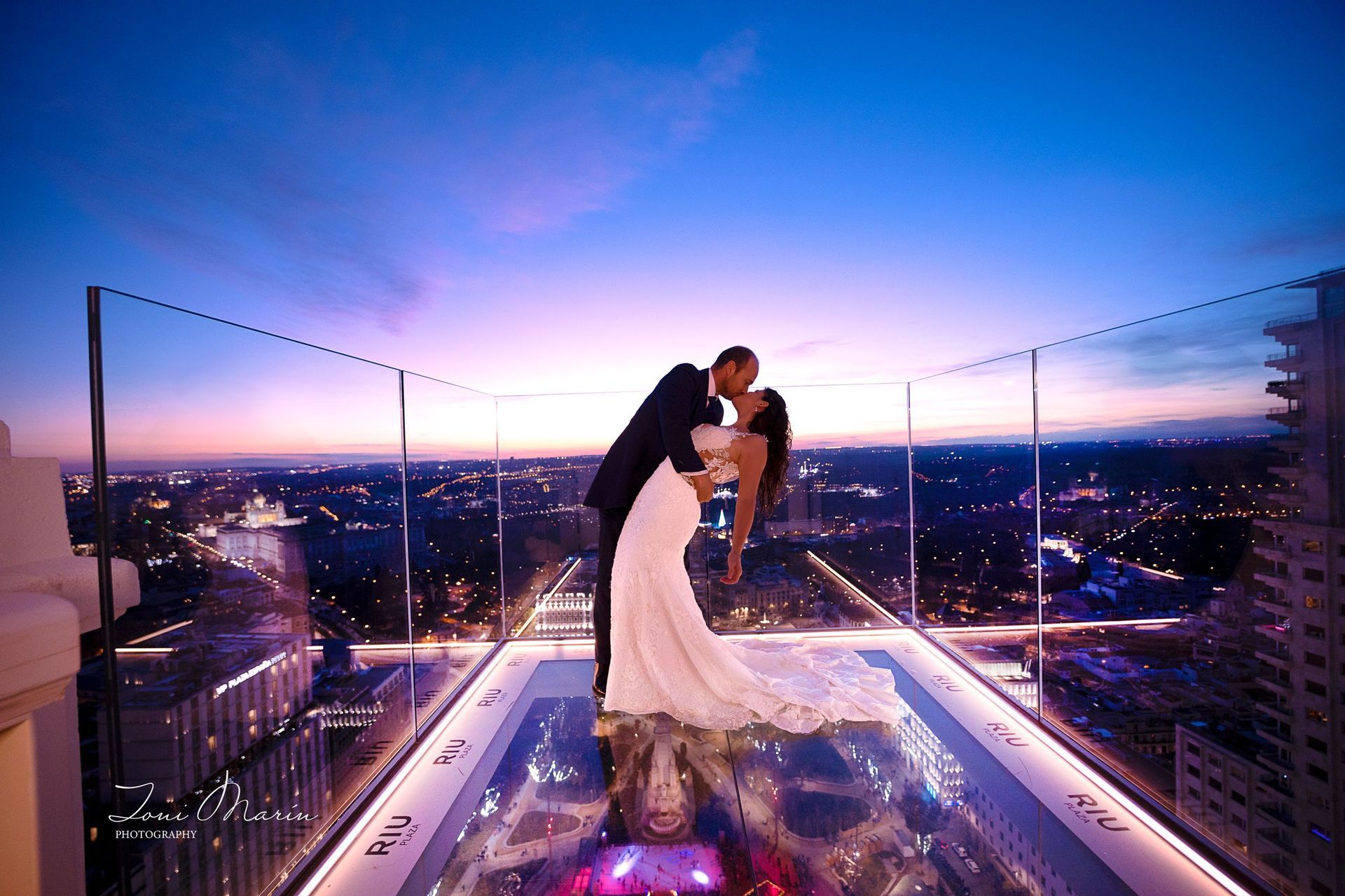 Una novia y un novio besándose en un balcón con vistas a una ciudad al atardecer.