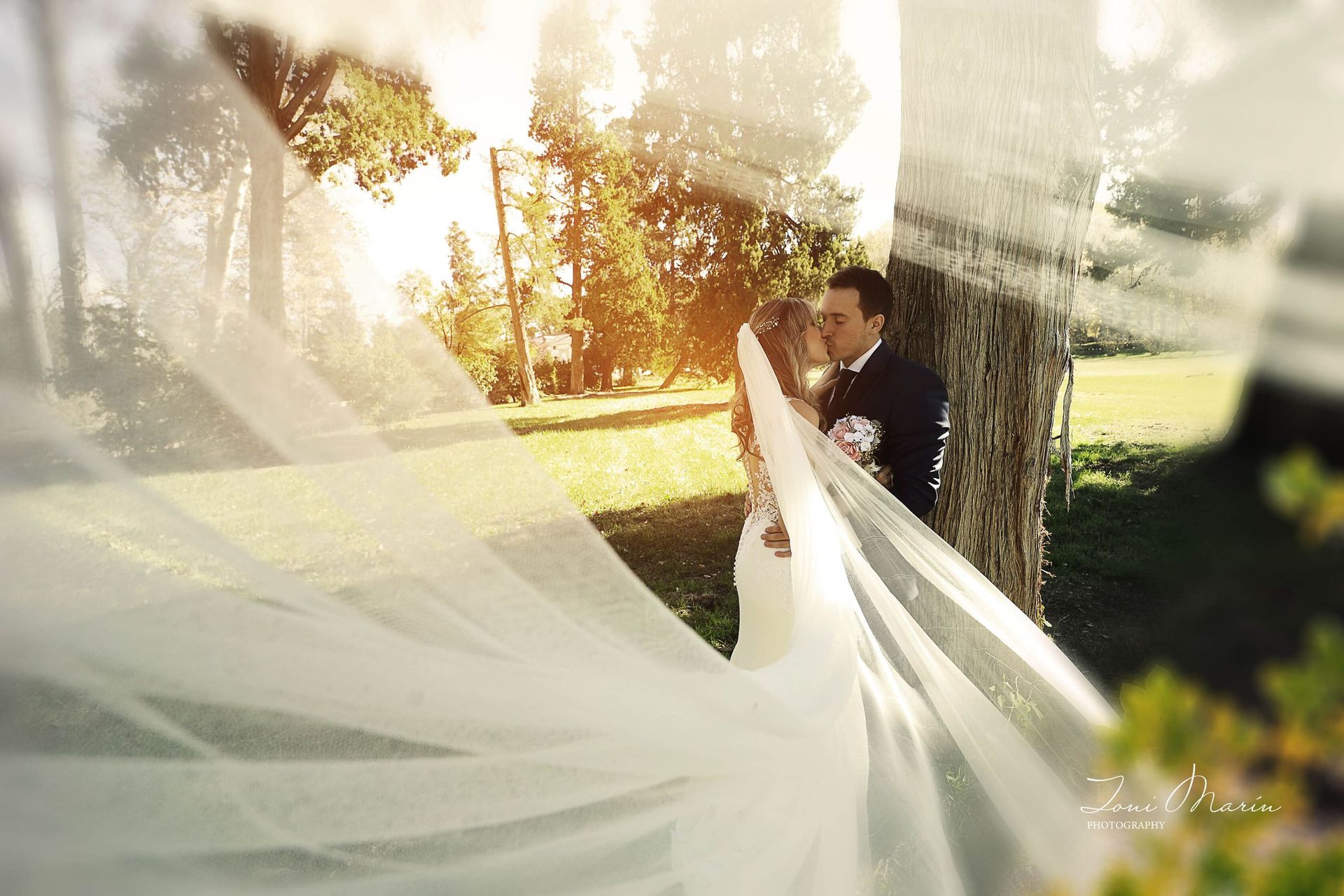 Una novia y un novio se besan bajo un velo que ondea al viento.
