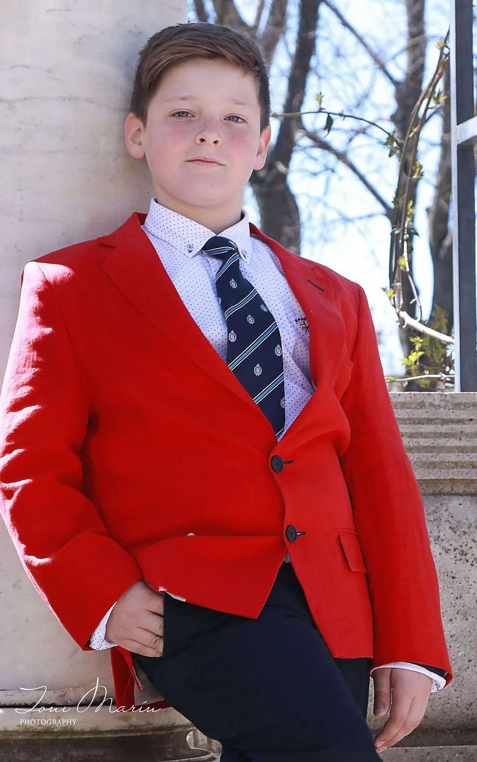 Un niño pequeño que lleva una chaqueta roja y corbata está apoyado contra una pared.