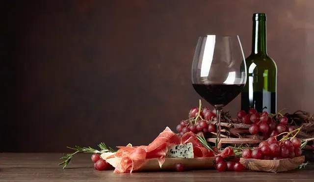 Una botella de vino, una copa de vino, uvas, jamón y queso sobre una mesa de madera.