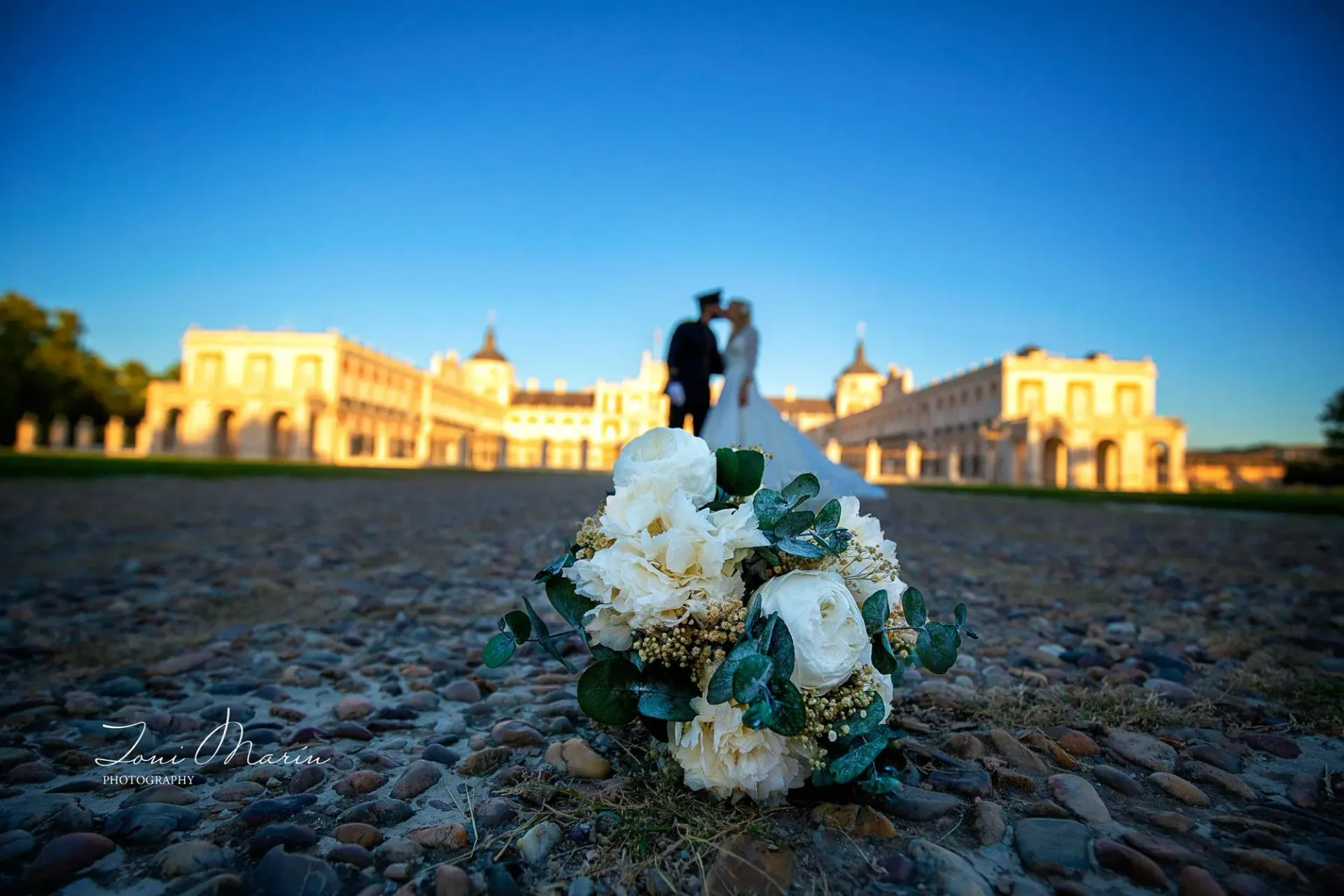 Una novia y un novio besándose frente a un edificio con un ramo de flores en primer plano.