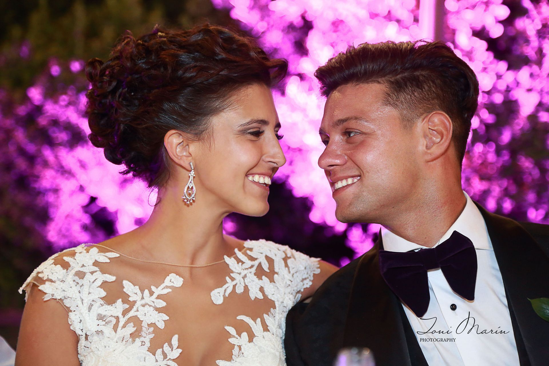 Una novia y un novio se sonríen delante de luces violetas.