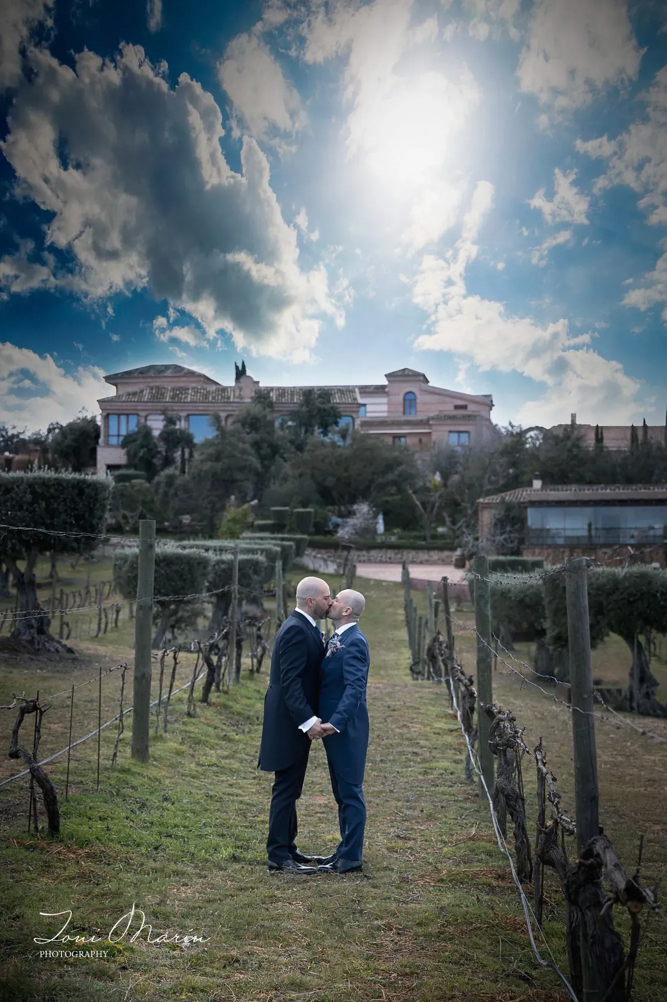 Dos hombres se besan en un campo con una casa al fondo.