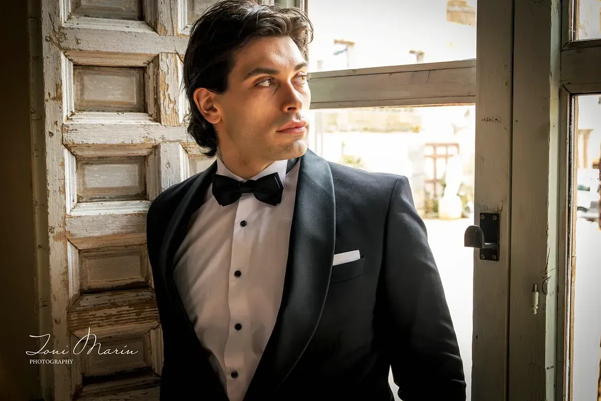 Un hombre con esmoquin y pajarita está de pie frente a una ventana.