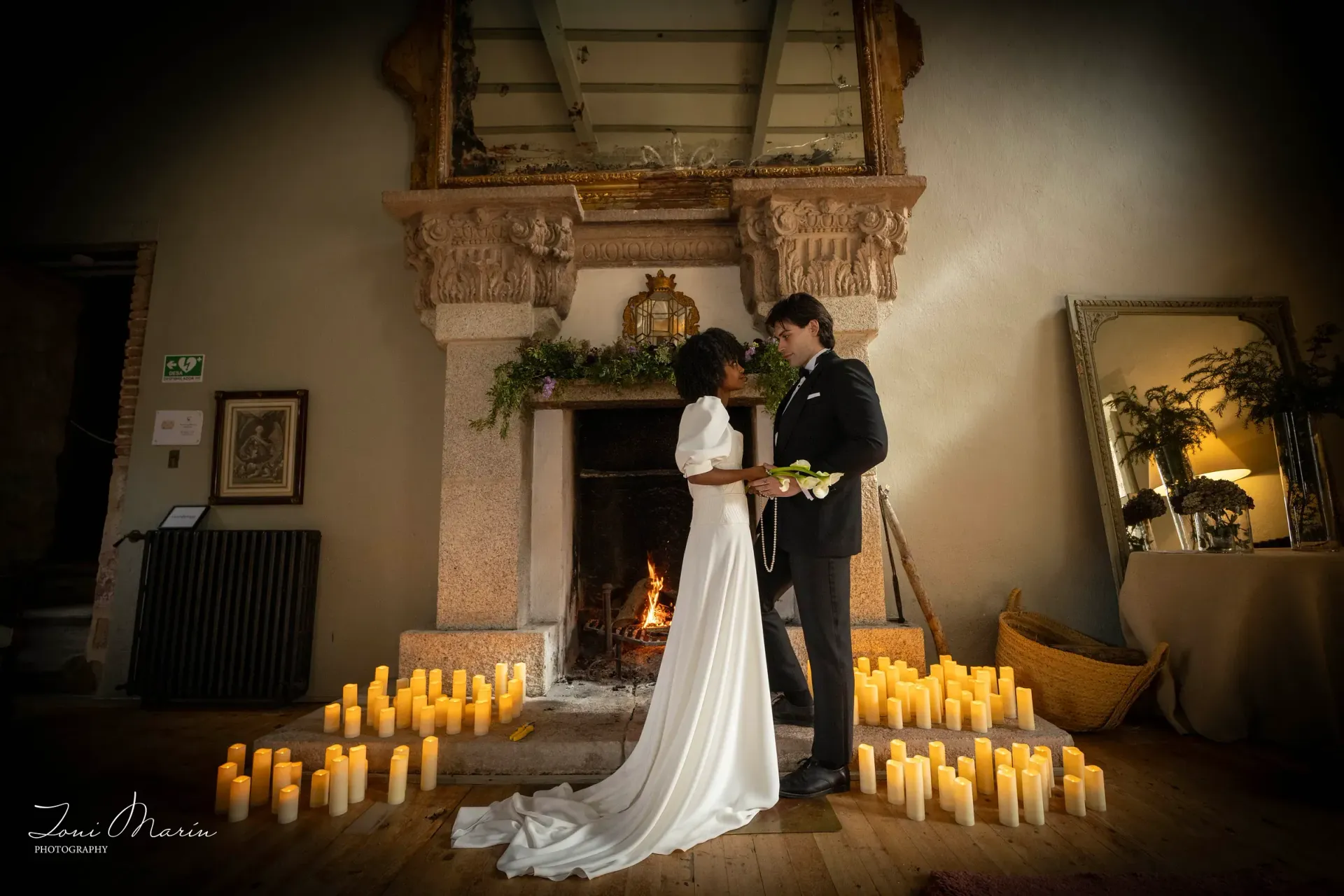 Una novia y un novio están de pie frente a una chimenea rodeados de velas.