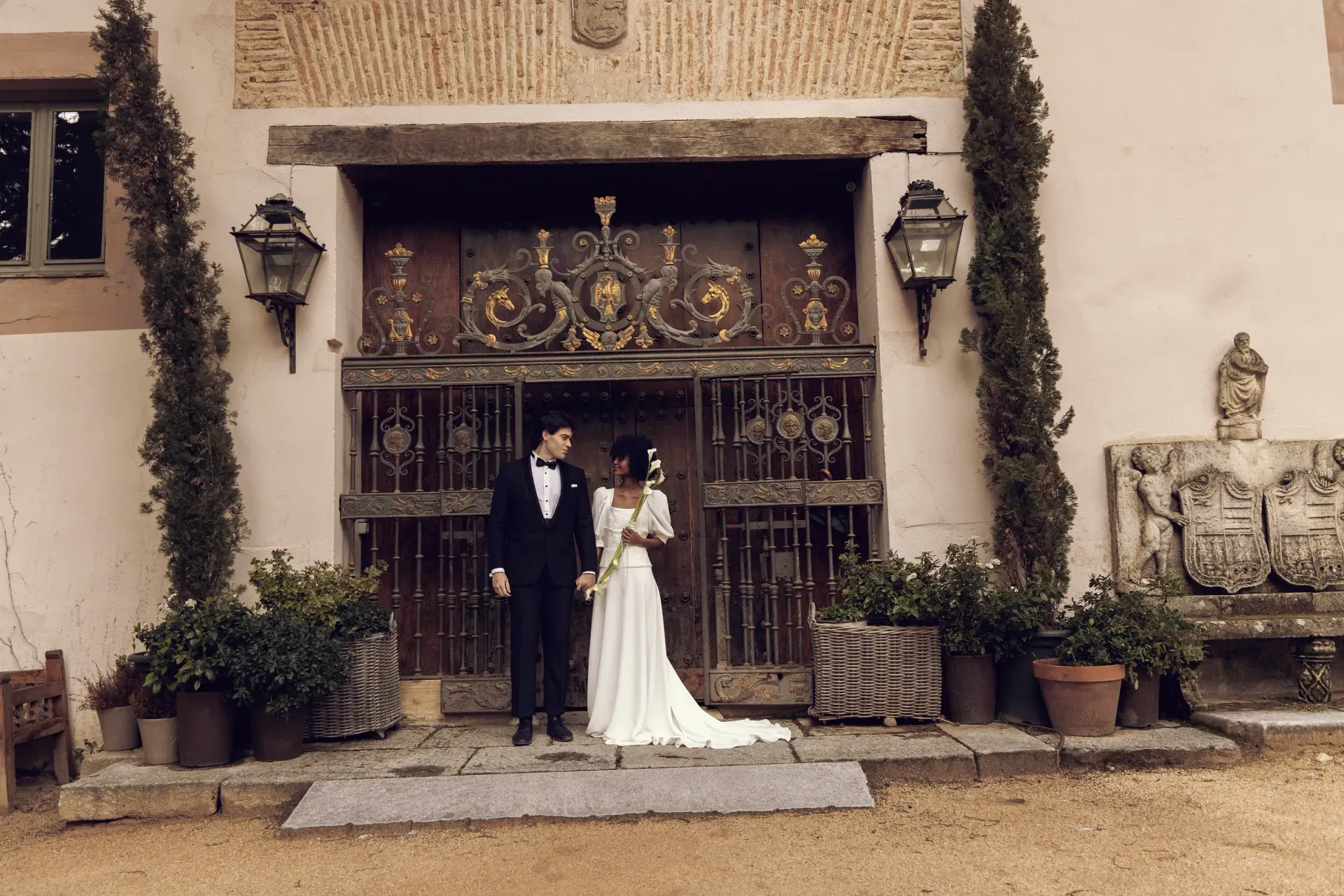 Una novia y un novio están parados frente a un edificio tomados de la mano.