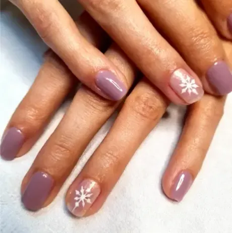 Manos con uñas cortas y redondeadas pintadas de color malva, con detalles de copos de nieve blancos en dos de ellas.