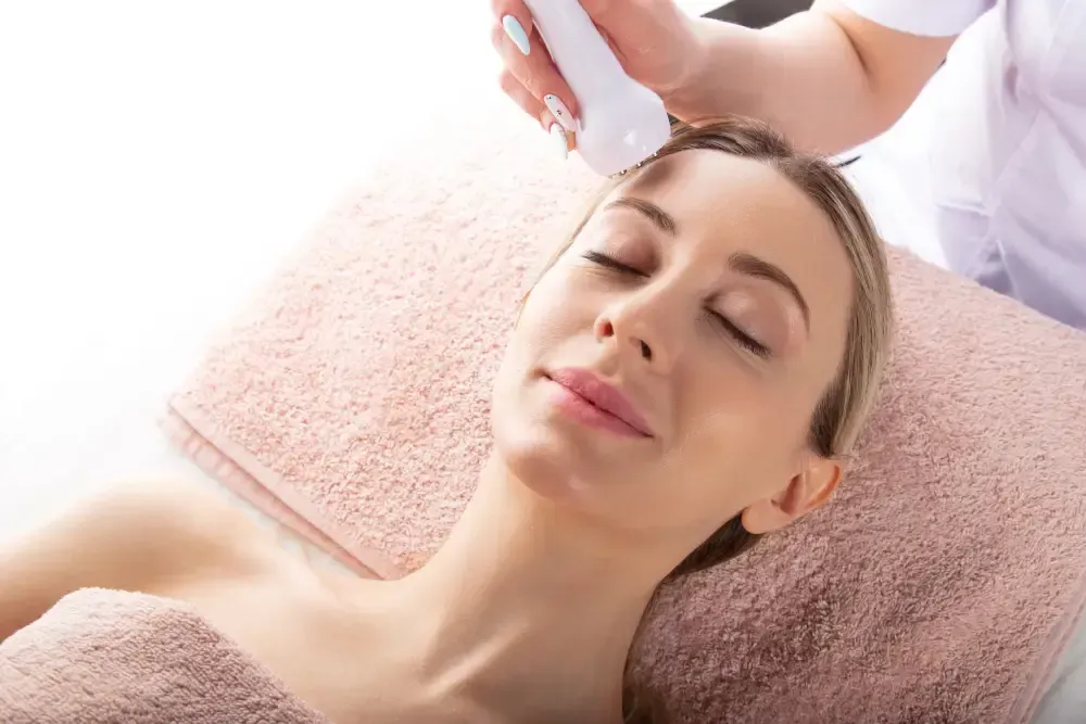 Una persona tumbada sobre una toalla suave mientras recibe un tratamiento facial con un dispositivo de mano.