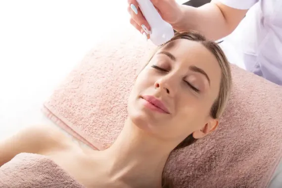 Una persona tumbada sobre una toalla suave mientras recibe un tratamiento facial con un dispositivo de mano.