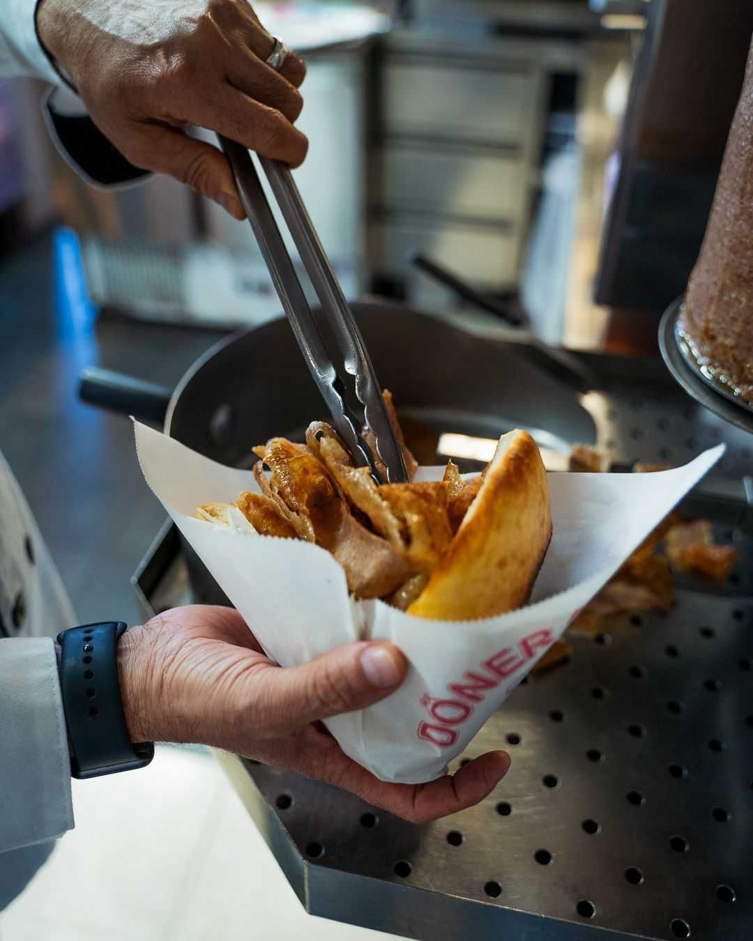 Ein Koch bereitet ein Döner-Kebab-Sandwich zu. Das Fleisch wird mit einer Zange in eine Brottüte gelegt.