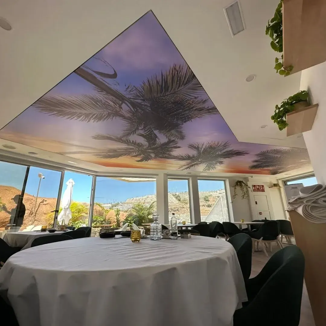 Interior de restaurante con mesas redondas dispuestas para cenar; mural de palmeras en el techo, vista de las montañas a través de las ventanas.