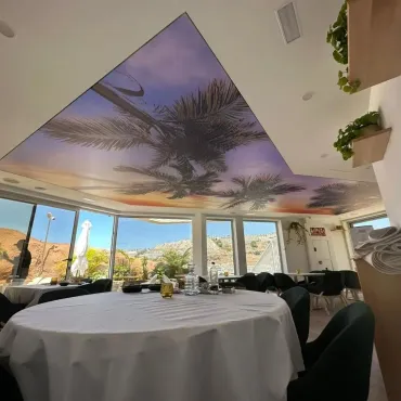 Interior de restaurante con mesas redondas dispuestas para cenar; mural de palmeras en el techo, vista de las montañas a través de las ventanas.