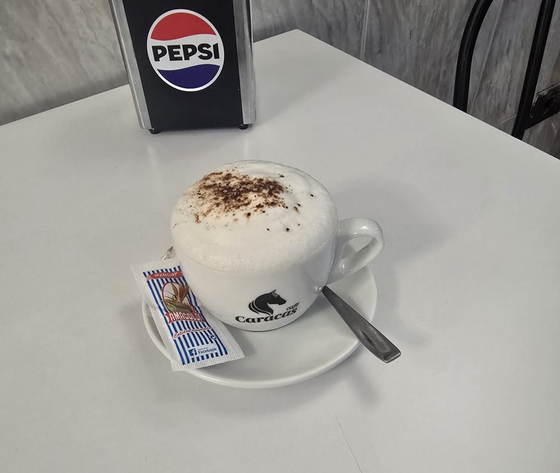 Taza de café batido sobre un platillo con cuchara y sobre de azúcar sobre una mesa, letrero de Pepsi al fondo.