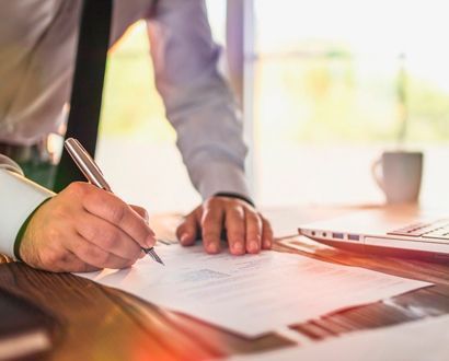 Persona firmando un documento en un escritorio, con camisa y corbata. Al fondo, una laptop y una taza de café.