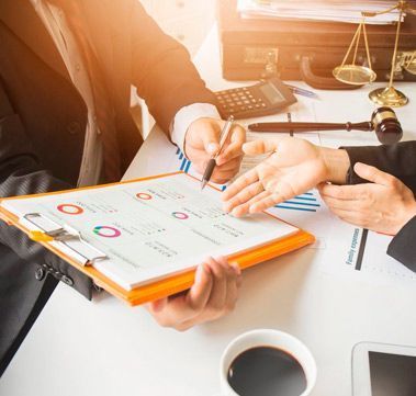 Dos personas trajeadas revisan informes financieros. Una señala con un bolígrafo; la balanza de la justicia al fondo.