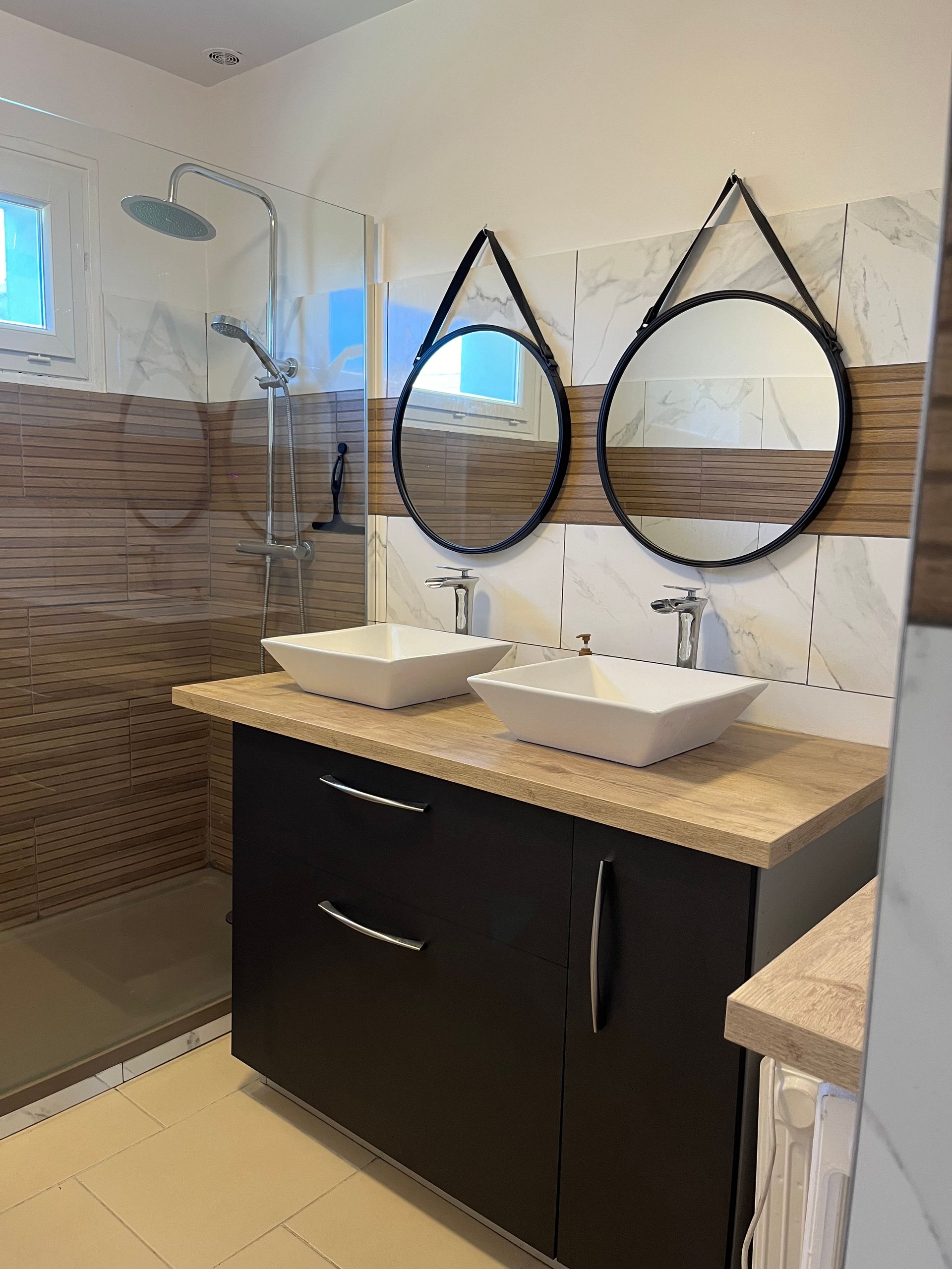 Meuble-lavabo moderne avec double vasque, miroirs ronds, douche carrelée et paroi de douche en verre.