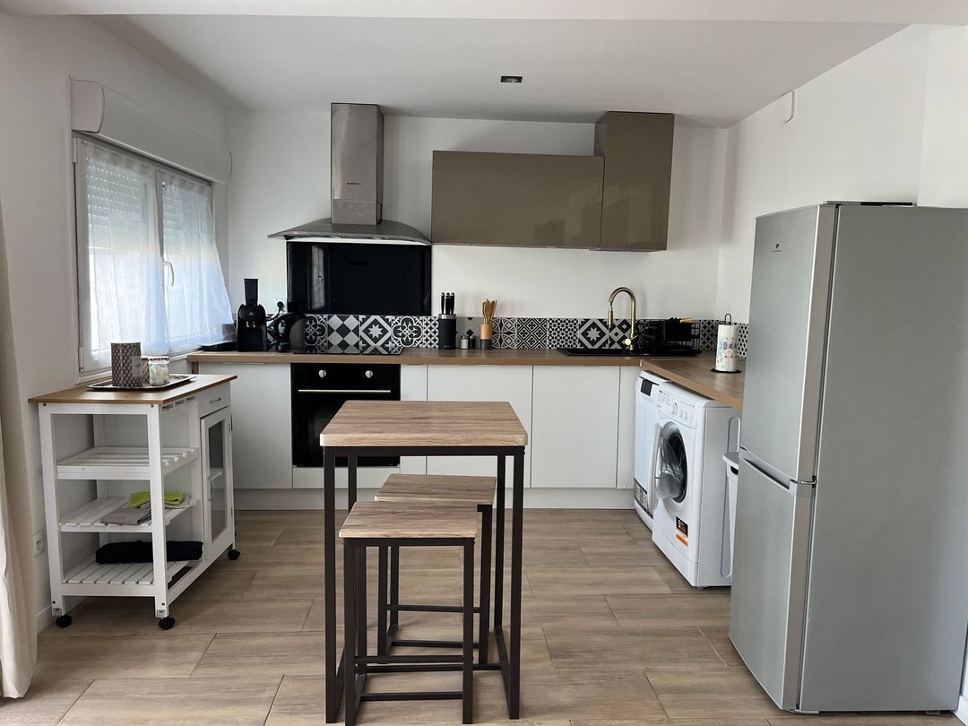 Cuisine moderne et lumineuse avec placards blancs, plans de travail en bois, une table et un grand réfrigérateur argenté.