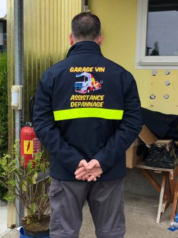Un homme en veste bleue avec l'inscription GARAGE VIN ASSISTANCE DÉPANNAGE dans le dos, se tient près d'un bâtiment.