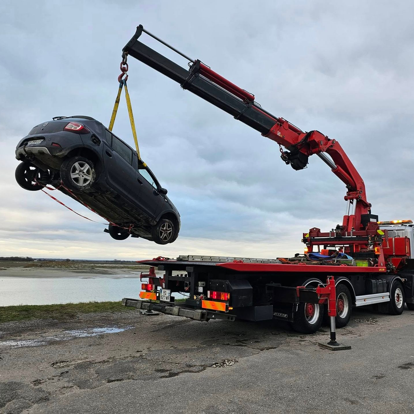 Une dépanneuse soulève un SUV foncé à l'aide d'une grue rouge près d'un point d'eau.