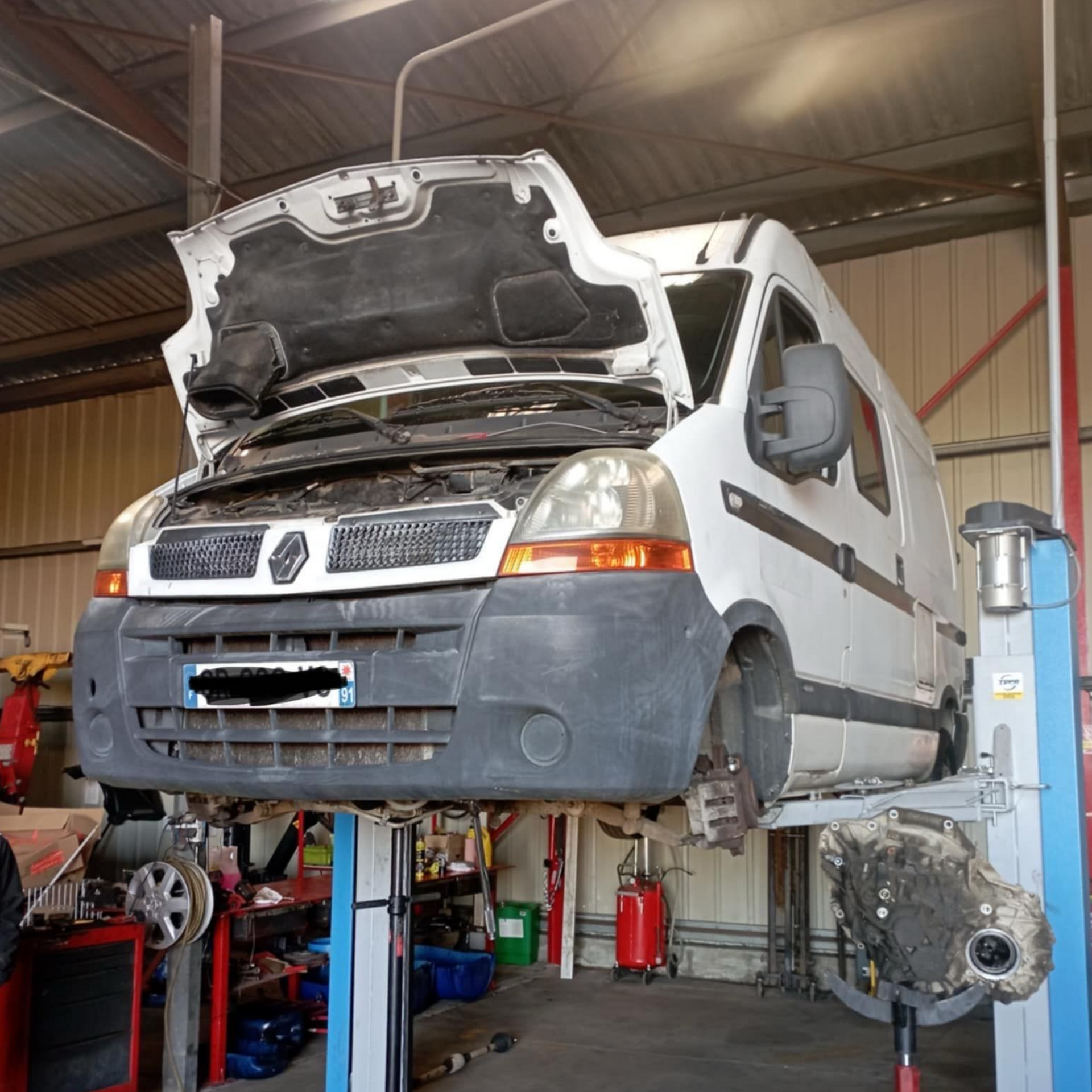 Fourgonnette blanche sur un pont élévateur, capot ouvert, dans un atelier de réparation.