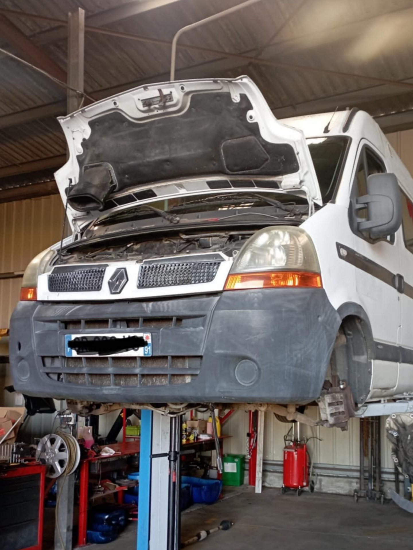 Fourgonnette blanche, capot ouvert, sur un pont élévateur dans un garage.