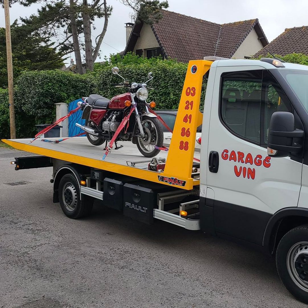 Moto sur une dépanneuse, moto rouge solidement attachée. La dépanneuse est blanche et porte l'inscription GARAGE VIN suivie d'un numéro.