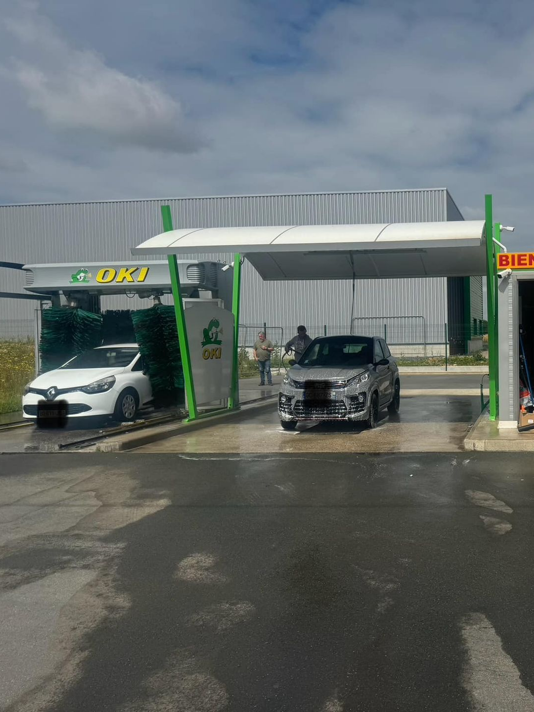 Deux voitures au lavage, une blanche, une argentée. Structure de lavage verte et blanche avec une personne à proximité.