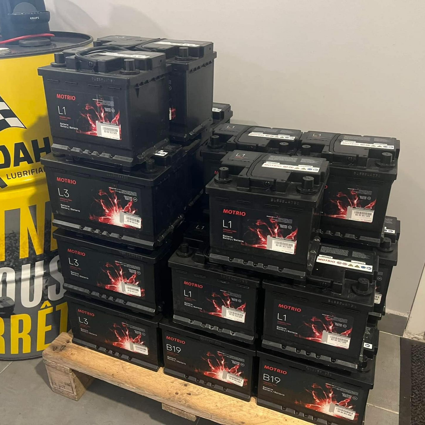 Une pile de batteries de voiture noires sur une palette en bois, à côté d'un fût jaune.